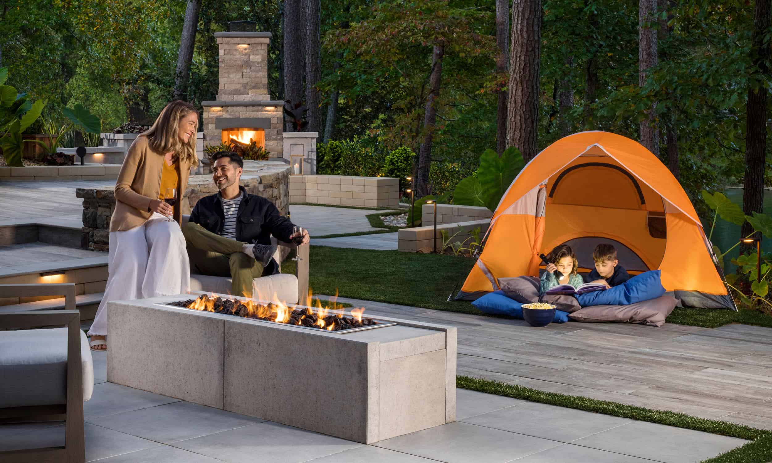 Family enjoying their Belgard outdoor living space. Mother and father sitting next to a fire pit while kids enjoy reading a book in a tent.