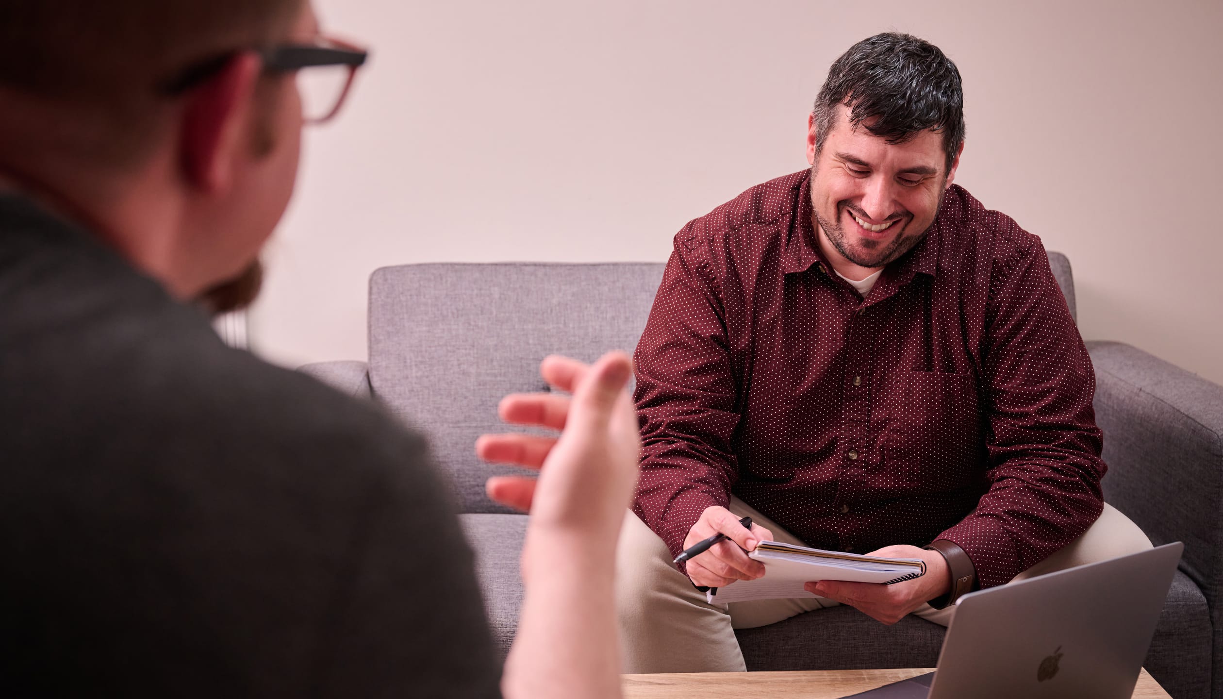 A man in a red shirt looking at a notebook, a man wearing glasses is out of focus in the foreground speaking to him